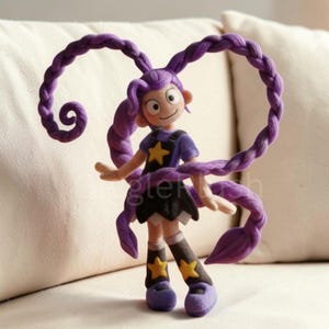 Pu&ograve; includere: Bambola fatta a mano in feltro con lunghi capelli viola intrecciati. La bambola indossa una maglietta viola con una stella gialla, una gonna nera e scarpe viola. I calzini della bambola hanno stelle gialle.
