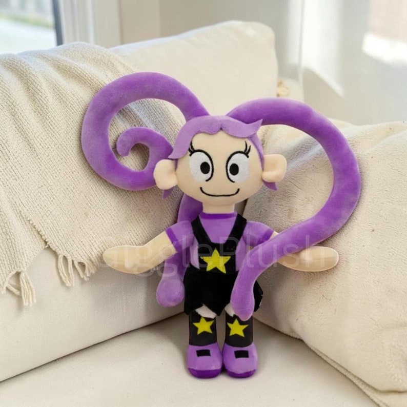 Pu&ograve; includere: Bambola di peluche viola con un viso sorridente, capelli viola e lunghi viticci ricci. Indossa una maglietta viola, una tuta nera con stelle gialle e stivali viola. La bambola &egrave; su un divano bianco.