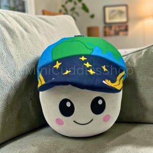 Puede incluir: Un peluche con forma de cara sonriente, que lleva una gorra azul y verde con un mapa del mundo y estrellas amarillas. Tiene ojos negros y mejillas rosas. El texto "MiniCuddlesShop" está en la gorra.