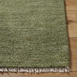 May include: A rectangular area rug in a solid olive green color featuring a dense, textured loop pile construction. The rug is finished with a short, straight fringe of off-white, uncolored fibers along the bottom edge of the textile piece.