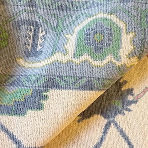 May include: Close-up of a section of a patterned rug. The rug features a light blue background with green and white floral designs. The rug is folded over, revealing a cream-colored underside.