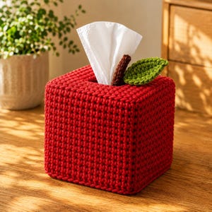 May include: A square tissue box cover crocheted in bright red yarn, featuring a small brown crocheted stem and a single green crocheted leaf attached to the top. A white tissue is pulled through the center opening of the apple-shaped cover.