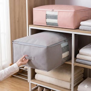 Puede incluir: Dos contenedores de almacenamiento, uno gris y otro rosa, están apilados en un estante. Cada contenedor tiene una cremallera en la parte superior y una ventana transparente que muestra toallas dobladas. Una persona saca el contenedor gris del estante.