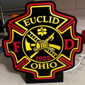 May include: A black, red, and yellow Maltese cross-shaped sign featuring the text EUCLID, F, D, OHIO, and 1922. The center displays a graphic of a firefighter helmet, a ladder, an axe, and a Star of Life symbol in a bold, high-contrast design.