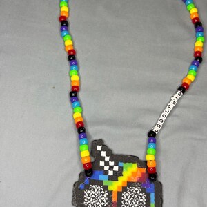 May include: A colorful beaded necklace with a pixelated design. The necklace features a rainbow of beads and a pendant with a black outline and a checkered pattern. The text "LSDOLPHIN" is spelled out in white beads.