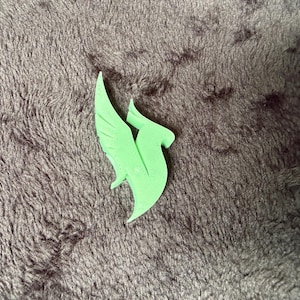 May include: A light green, flat, stylized emblem shaped like a bird or phoenix in flight with pointed wings and a curved tail. The object features a smooth, matte surface and sharp, angular edges, depicting a singular, abstract avian silhouette.