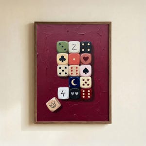 May include: A framed wall art piece featuring a deep red textured background with a grid of thirteen square tiles displaying various symbols including numbers, card suits, a moon, and hearts, plus one loose tile with a crown icon at the bottom left.