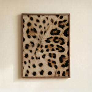 May include: A framed print featuring a close-up of a leopard print pattern. The design showcases a beige background with black and brown spots, framed in a dark brown wooden frame. The print is rectangular and mounted on a light beige wall.