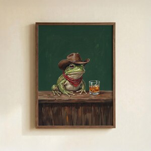 May include: A framed art print featuring a green frog wearing a brown cowboy hat and a red bandana. The frog sits at a wooden bar with a glass of whiskey. The background is a dark green color.
