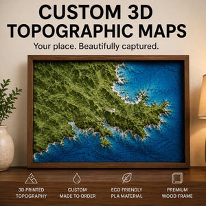 May include: A rectangular 3D topographic map in a dark wood frame, displaying a green coastal landscape meeting deep blue water. Text above reads CUSTOM 3D TOPOGRAPHIC MAPS. Your place. Beautifully captured. Icons below note 3D printed topography.