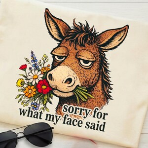 May include: Cream-colored t-shirt featuring a cartoon donkey with a bouquet of flowers in its mouth. The donkey has a slightly grumpy expression. Text on the shirt reads "sorry for what my face said". Black sunglasses are in the foreground.