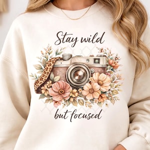May include: Cream-colored sweatshirt featuring a watercolor design of a vintage camera surrounded by flowers. The text "Stay wild but focused" is above and below the camera. The camera strap has a leopard print.