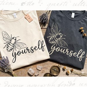 May include: Two t-shirts, one cream and one charcoal, each with a large bee graphic and the word "yourself" in a script font. The cream shirt has a Boho Bee tag. The shirts are on a wooden surface with lavender and other decorative items.