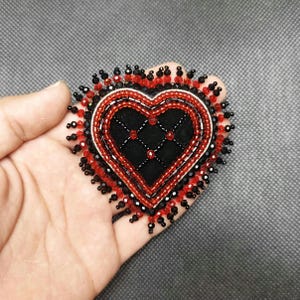 May include: A heart-shaped brooch with a black velvet center, adorned with red and black beads. The brooch features a quilted pattern in the center and is outlined with red and black beads. The edges are finished with a beaded fringe.
