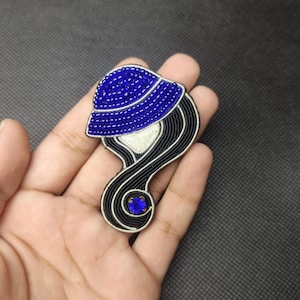 May include: A brooch shaped like a stylized head wearing a blue beaded hat. The hair is formed from black cord with silver metallic outlines. A single round blue rhinestone is set at the base of the hair, and the face area is a smooth cream color.