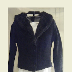 May include: A black cardigan sweater with a faux fur collar. The sweater has long sleeves and a button-up front. The collar is a dark, textured material. The sweater is displayed on a wire dress form, with a white shirt underneath.