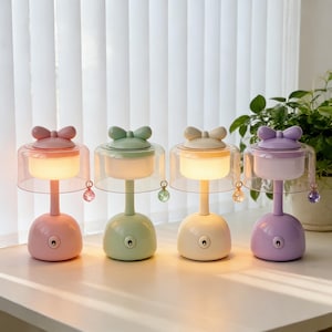 May include: Four pastel-colored decorative lamps with a bow-shaped top and a clear, bell-shaped shade. Each lamp has a different color: pink, green, cream, and purple. A crystal charm hangs from each shade. The lamps are on a white surface.