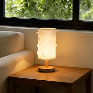 Puede incluir: Lámpara de mesa decorativa con una pantalla cilíndrica blanca adornada con recortes de mariposas. La lámpara tiene una base y un tallo de madera, colocada sobre una mesa de madera. La lámpara está encendida, emitiendo una luz cálida.