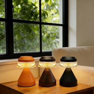 Könnte beinhalten: Drei moderne Tischlampen mit konischen Sockeln in Orange, Braun und Schwarz. Jede Lampe hat einen dekorativen, transparenten, gemusterten Schirm und strahlt ein warmes Licht aus. Die Lampen stehen auf einer Holzoberfläche in der Nähe eines Fensters.