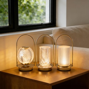 Op de afbeelding: Drie decoratieve tafellampen met goudkleurige metalen frames en bases. Elke lamp heeft een andere textuur glazen kap: een bol, een cilinder met een kruispatroon en een gegroefde cilinder. De lampen zijn verlicht en geven een warme gloed.