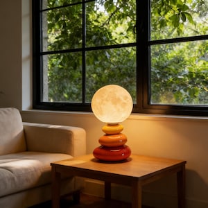 Puede incluir: Lámpara de mesa decorativa con una luz en forma de luna y una base de piedras apiladas. La lámpara emite un brillo cálido, con una luz esférica en la parte superior y una base de piedras naranjas, amarillas y rojas. La lámpara está sobre una mesa de madera.