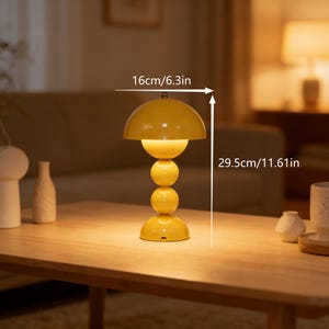 May include: A yellow table lamp with a mushroom-shaped shade and a stacked sphere base. The lamp is 11.61 inches tall and 6.3 inches wide. The lamp is on a wooden table. The lamp is turned on, emitting a warm glow.