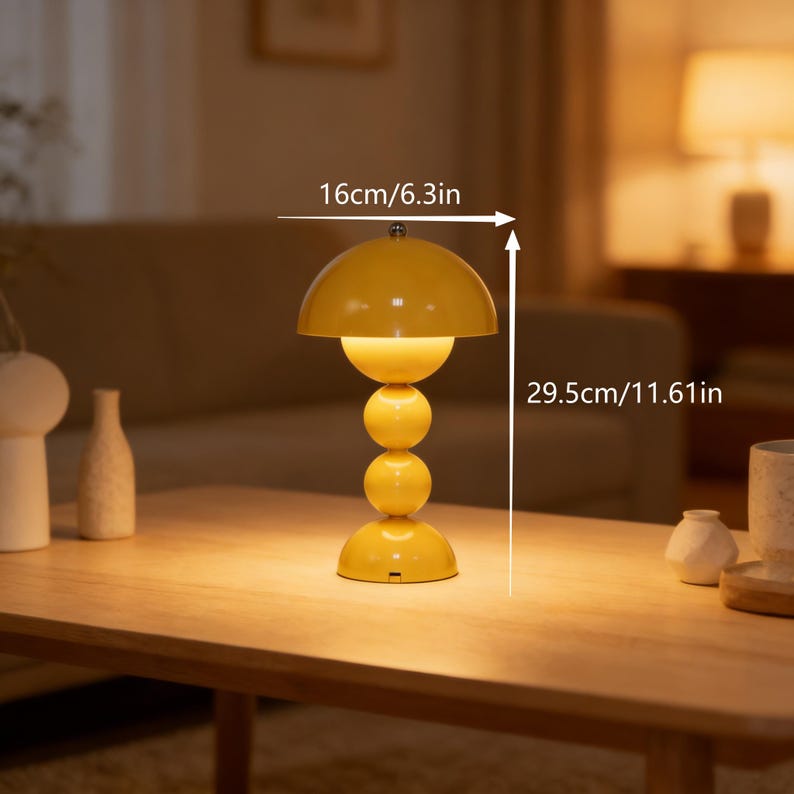 May include: A yellow table lamp with a mushroom-shaped shade and a stacked sphere base. The lamp is 11.61 inches tall and 6.3 inches wide. The lamp is on a wooden table. The lamp is turned on, emitting a warm glow.