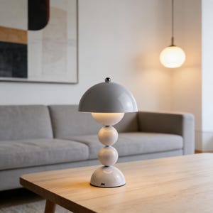 May include: A modern table lamp with a gray, dome-shaped shade and a white, spherical base. The lamp has a minimalist design and sits on a wooden table. The lamp's design is reminiscent of a mushroom.