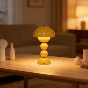 May include: A yellow table lamp with a rounded shade and a stacked sphere design. The lamp is made of metal and sits on a wooden table. The lamp is turned on, emitting a warm glow.