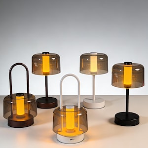 May include: A collection of modern, battery-powered lamps with a cylindrical design. Each lamp features a warm, amber-colored light encased in a clear, dome-shaped shade. The lamps have bases and stems in various colors, including black, white, and brown.