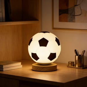 May include: A soccer ball-shaped lamp with black pentagons and white panels, resting on a round wooden base. The lamp is illuminated, casting a warm glow. The lamp is on a wooden desk with books and a pen holder.