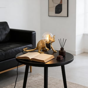 May include: A gold-coloured cat-shaped table lamp with a round light bulb. The lamp is on a black round table with an open book, a diffuser, and a small chocolate. The lamp's cord is visible.