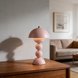 May include: A pink table lamp with a rounded shade and a stacked sphere base. The lamp has a silver-colored top and is placed on a wooden surface. The lamp's design is modern and decorative, suitable for home decor.