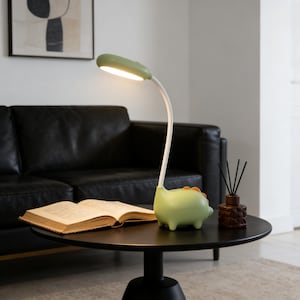 Puede incluir: Una lámpara de escritorio verde y blanca con diseño de dinosaurio. La lámpara tiene un cuello flexible y una pantalla verde claro. La base de la lámpara tiene forma de dinosaurio con detalles en naranja. Un libro abierto está sobre una mesa negra.
