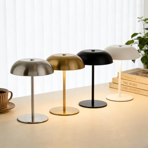 Puede incluir: Cuatro lámparas de mesa modernas con una pantalla en forma de cúpula y un tallo delgado. Las lámparas son de color plateado, dorado, negro y blanco. Están dispuestas sobre una superficie clara, con una taza y libros en el fondo.