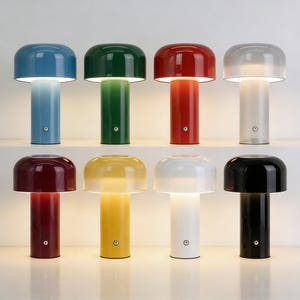 Op de afbeelding: Een verzameling van acht paddenstoelvormige tafellampen in verschillende kleuren, waaronder blauw, groen, rood, grijs, bordeaux, geel, wit en zwart. Elke lamp heeft een afgeronde kap en een cilindrische voet, met een zachte gloed die uit de onderkant van de kap komt.