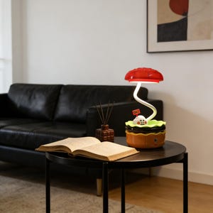 May include: A whimsical mushroom-shaped table lamp with a red cap and a white, curved stem. The lamp features a small figure with a "SALE" sign. The lamp sits on a round, black table next to an open book.
