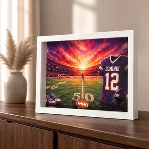 Könnte beinhalten: Ein weiß gerahmter Druck eines Footballstadions bei Sonnenuntergang mit einem violetten Trikot mit der Aufschrift DOMINIK und der Nummer 12, einem Football und Stollen auf dem Spielfeld unter einem leuchtend orange-violetten Himmel.
