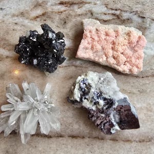 May include: Four different mineral specimens are displayed on a marble surface. The minerals include a cluster of black crystals, a pink, textured rock, a clear quartz formation, and a gray and black mineral.