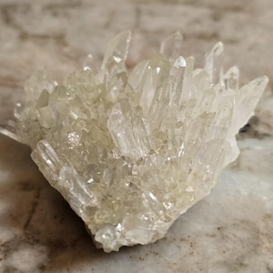 May include: A cluster of clear quartz crystals with sharp, pointed terminations. The crystals are translucent and arranged in a dense, irregular formation. The quartz has a slightly milky appearance and sits on a light-colored surface.