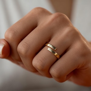 Peut inclure: Une bague enveloppante dorée avec les noms "ELIJAH" et "OLIVIA" gravés. La bague est portée sur une main, les noms étant affichés sur le doigt. La bague a un design simple et élégant.