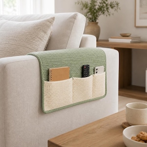 May include: A green and cream-colored crochet sofa arm organizer with three pockets. The organizer is draped over the arm of a light-colored sofa. A notebook, remote control, and smartphone are in the pockets.