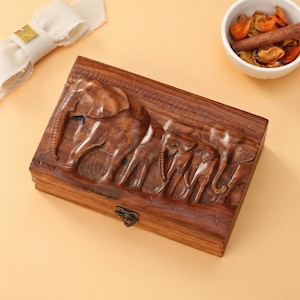May include: A rectangular wooden box with a carved relief of a herd of elephants. The box is a warm brown color and has a metal clasp. The elephants are intricately detailed, with visible tusks and wrinkled skin. The box is likely used for storage.