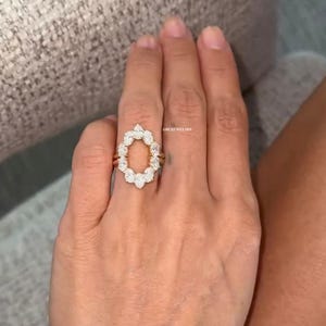 May include: A gold ring featuring an oval design of white gemstones. The ring has a halo of smaller stones around the oval, with a delicate band. The ring is on a person's finger.