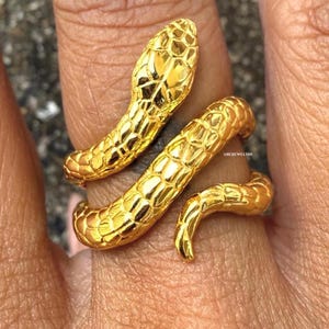 Peut inclure: Une bague serpent dorée avec un motif texturé en forme d'écailles. La tête du serpent est relevée et le corps s'enroule autour du doigt. La bague est en métal brillant et réfléchissant.