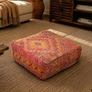 May include: A square, patterned floor pouf in shades of red, orange, and purple sits on a neutral-toned rug. The pouf has a geometric design. The room also features a beige sofa, wooden coffee table, and a wooden cabinet.