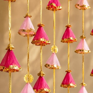 May include: A decorative hanging garland featuring multiple vertical gold ribbons adorned with alternating cone-shaped tassels in shades of hot pink and light pink. Each fabric tassel is finished with a metallic gold trim at the base and top.