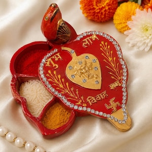 Puede incluir: Un recipiente rojo en forma de corazón con una tapa que presenta un emblema de deidad dorado, pedrería y texto en hindi. La base tiene tres compartimentos con polvo rojo, arroz blanco y cúrcuma amarilla, coronado por una figura de ave roja.