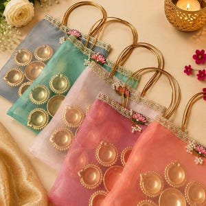 Puede incluir: Un conjunto de cinco bolsas de regalo de organza transparente en gris, verde azulado, rosa claro, magenta y coral, cada una con asas de alambre dorado, un borde de encaje metálico y un dije de flor de loto rosa con cuentas blancas y lámparas diya doradas.