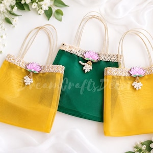 May include: Three decorative tote bags. Two are yellow, and one is green. Each bag has a gold handle, a pink lotus flower, and a white beaded accent. The bags have a lace trim along the top edge. The bags are on a white surface.